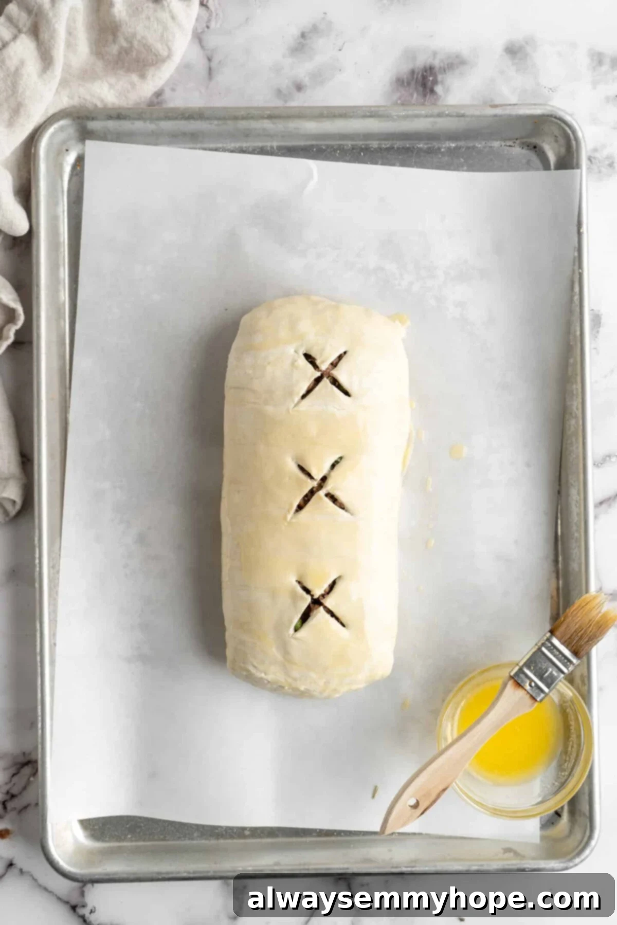 This vegan wellington recipe uses mushrooms, walnuts, and vegan beef to replace meat. It's a hearty holiday meal that's easy to make. An uncooked vegan wellington resting on parchment paper, perfectly shaped and ready for the oven, with a small bowl of melted vegan butter and a pastry brush poised nearby for the final glaze.