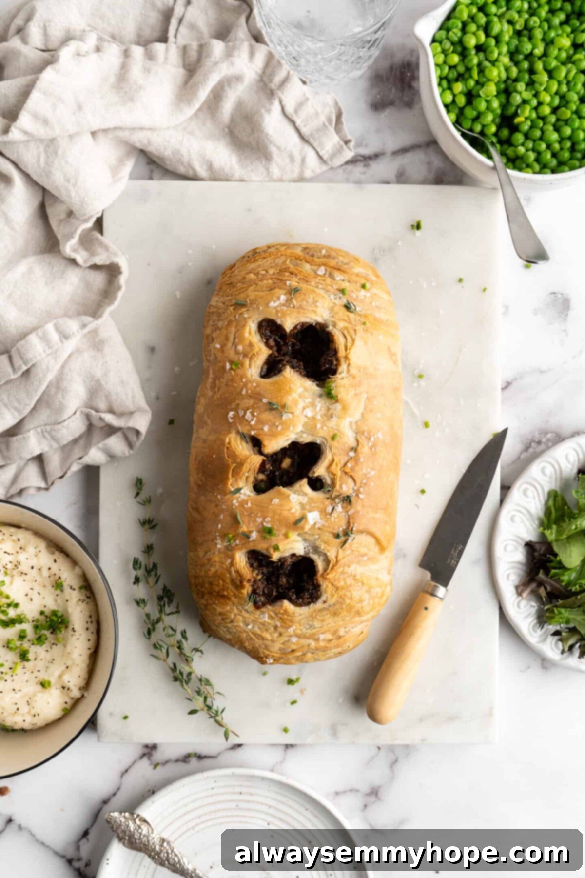 This vegan wellington recipe uses mushrooms, walnuts, and vegan beef to replace meat. It's a hearty holiday meal that's easy to make. A golden-brown vegan wellington with artfully scored marks on its top, presented on a wooden cutting board with a paring knife, fresh thyme sprigs, a kitchen towel, and delectable side dishes of mashed potatoes, peas, and a fresh green salad, forming a complete and inviting meal.