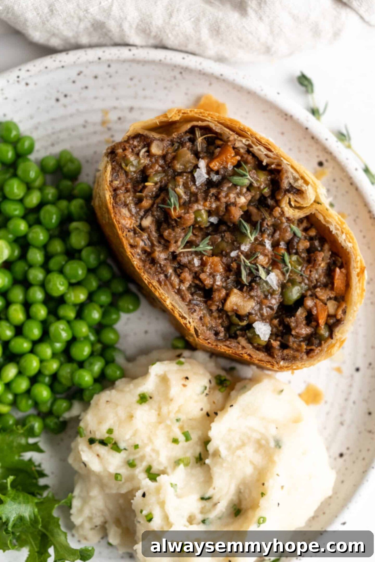 This vegan wellington recipe uses mushrooms, walnuts, and vegan beef to replace meat. It's a hearty holiday meal that's easy to make. A perfectly sliced portion of vegan wellington presented on a white plate, accompanied by a side of vibrant green peas and a mound of fluffy mashed potatoes, creating a complete and satisfying plant-based meal.
