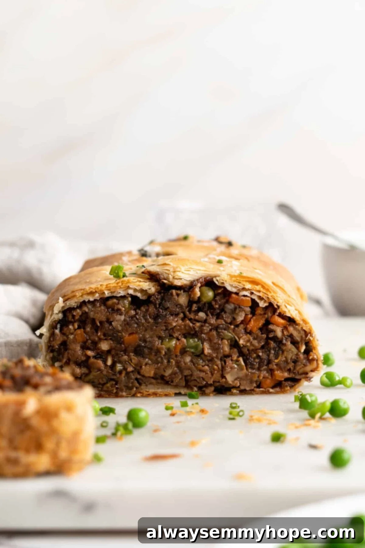 This vegan wellington recipe uses mushrooms, walnuts, and vegan beef to replace meat. It's a hearty holiday meal that's easy to make. A close-up, front-on view of a perfectly baked vegan wellington, sliced cleanly in half to showcase its rich, textured filling, beautifully surrounded by bright green peas on a white plate.