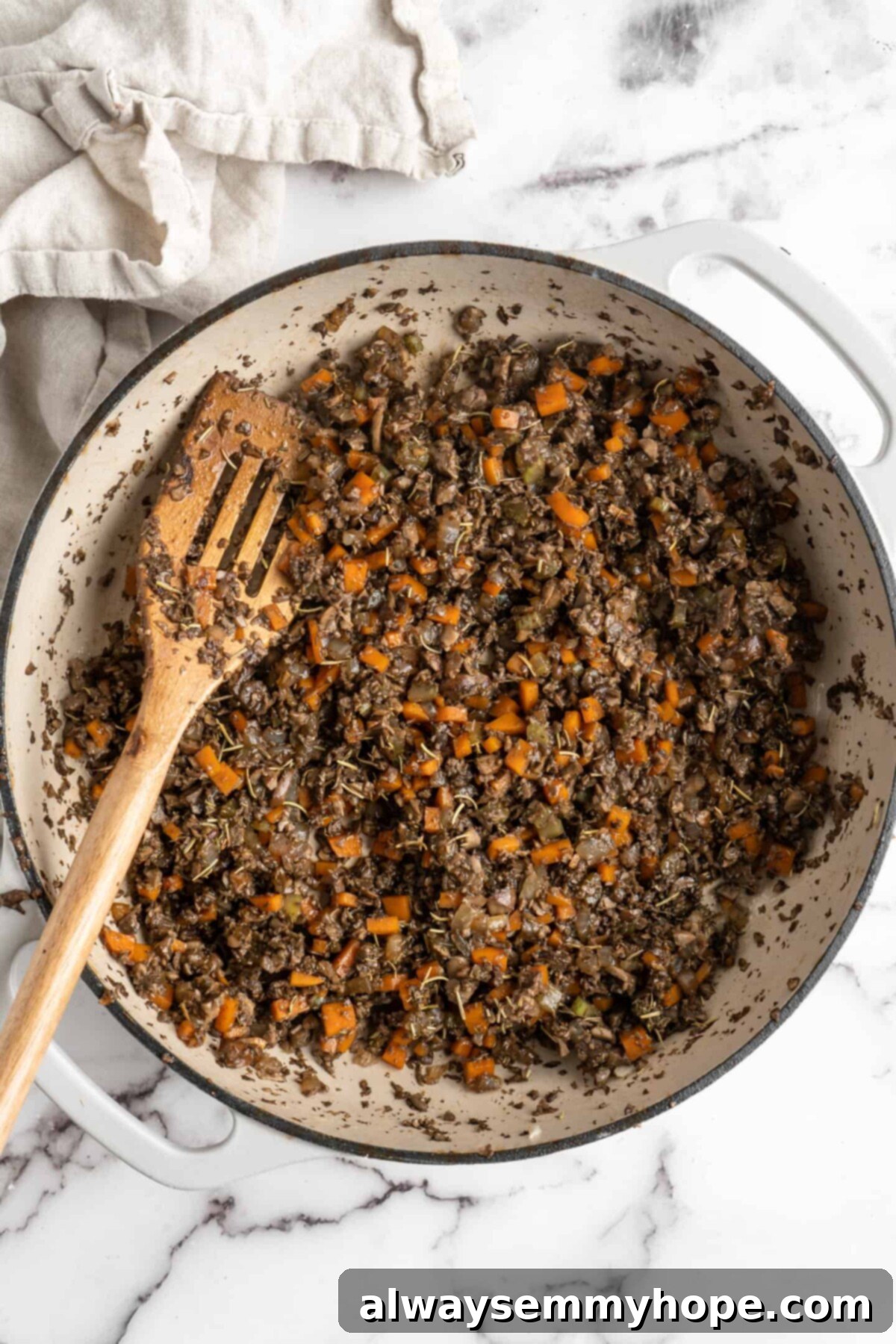 This vegan wellington recipe uses mushrooms, walnuts, and vegan beef to replace meat. It's a hearty holiday meal that's easy to make. A vibrant mix of diced mushrooms, carrots, onions, and celery gently cooking in a pan with a slotted wooden spoon, steam subtly rising, indicating the softening and release of liquid from the vegetables for the wellington filling.