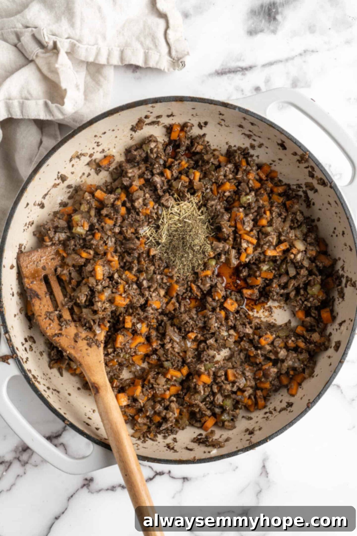 This vegan wellington recipe uses mushrooms, walnuts, and vegan beef to replace meat. It's a hearty holiday meal that's easy to make. A pan filled with sautéed mushrooms, onions, carrots, and celery, topped with a generous sprinkle of dried rosemary and thyme, with a wooden slotted spoon resting in the mix, capturing the aromatic stage of the vegan wellington filling preparation.