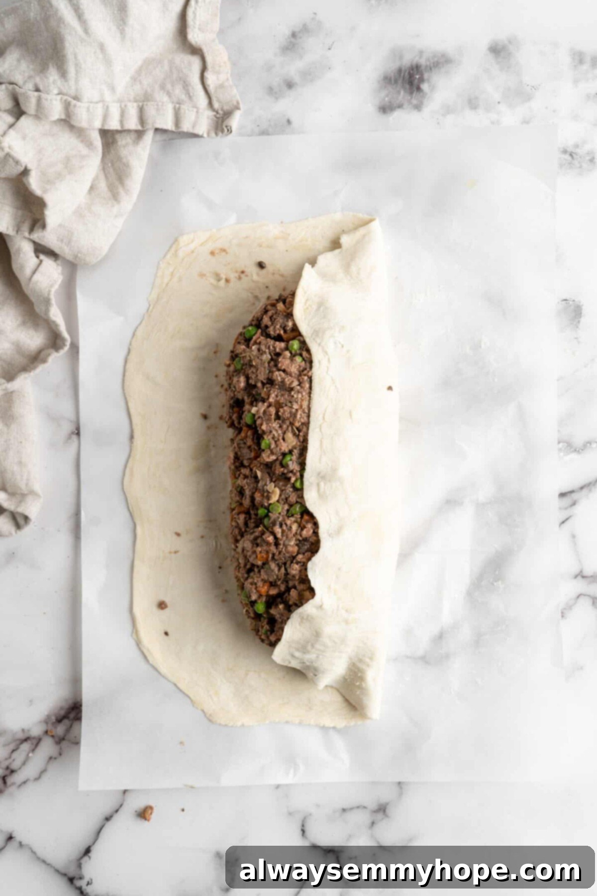 This vegan wellington recipe uses mushrooms, walnuts, and vegan beef to replace meat. It's a hearty holiday meal that's easy to make. A sheet of rolled-out puff pastry with a log of vegan wellington filling in the middle, showing one long end of the pastry neatly folded over the top of the filling, beginning the wrapping process.