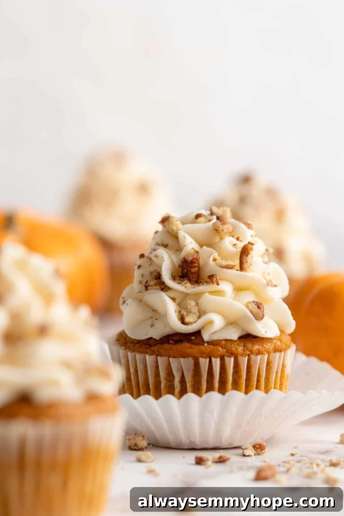 Close up of a vegan pumpkin cupcake topped generously with creamy vegan cream cheese frosting and chopped pecans, with more beautifully frosted cupcakes and decorative pumpkins softly blurred in the background. A single, perfectly baked cupcake sits enticingly in the foreground.