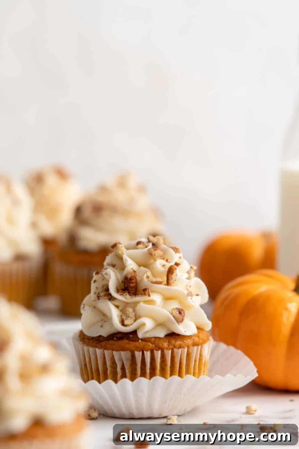 A single, perfectly baked vegan pumpkin cupcake topped with a swirl of rich cream cheese frosting and a sprinkle of pecans, set against a softly blurred background of more frosted cupcakes and decorative pumpkins.