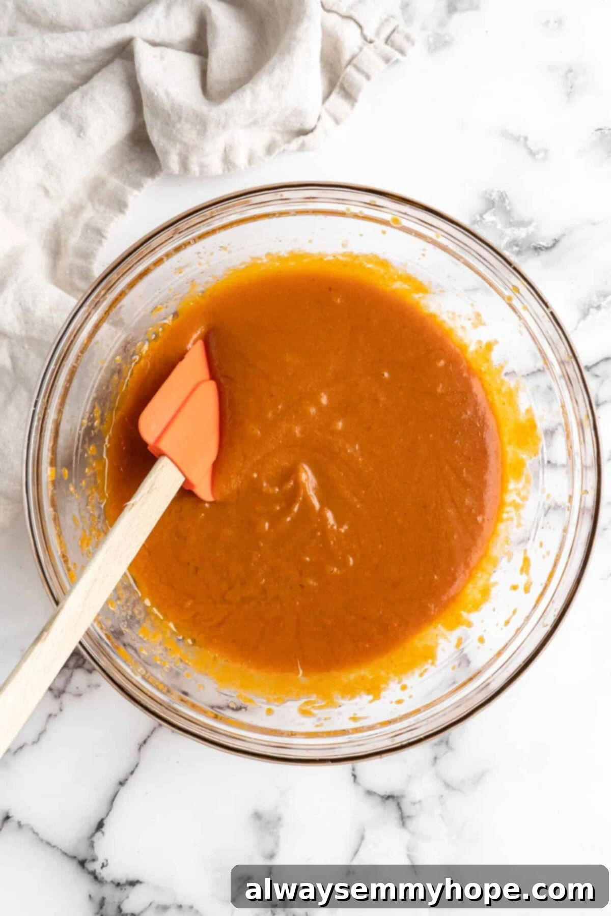 A mixing bowl holding a smooth, vibrant orange mixture of pumpkin puree, vegan milk, and sugar, thoroughly combined with a rubber spatula resting on the side.