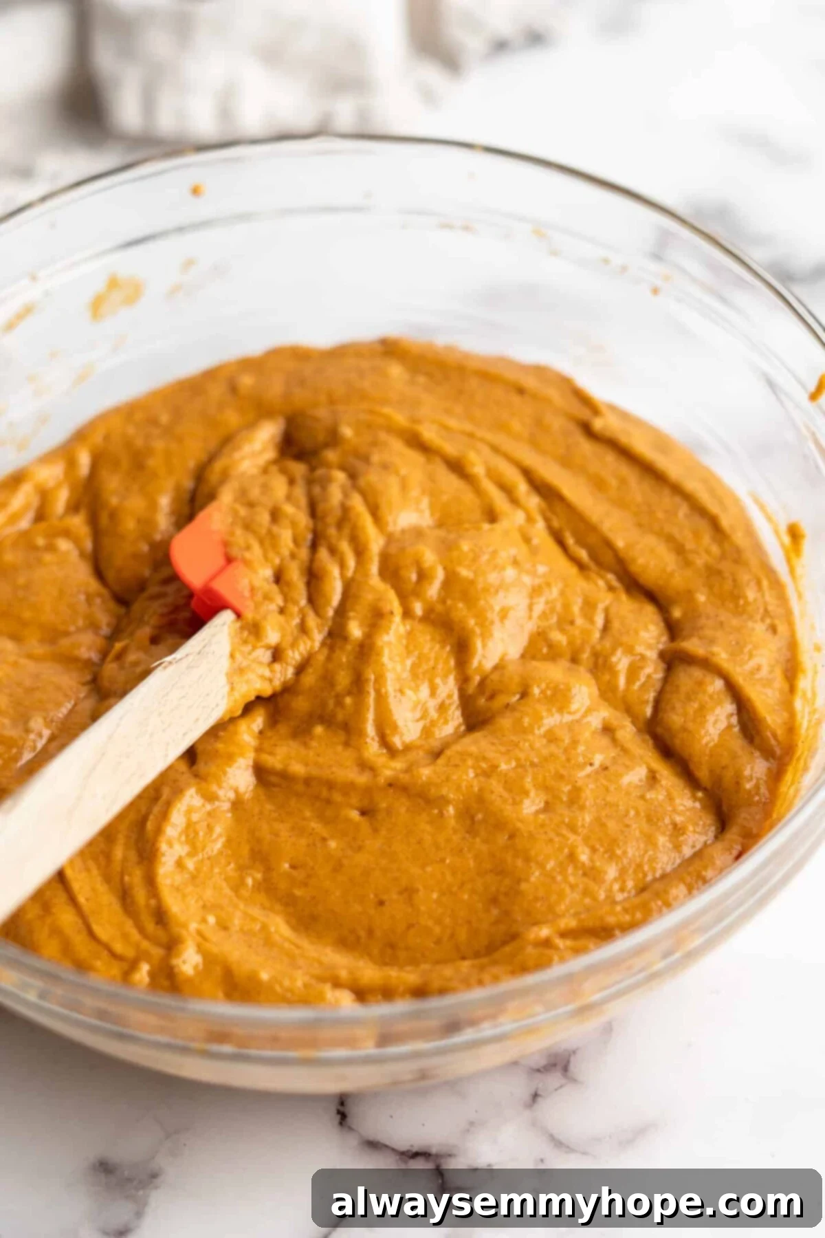 A mixing bowl overflowing with beautifully smooth pumpkin cupcake batter, a rubber spatula poised to scrape down the sides, showcasing its perfect consistency.