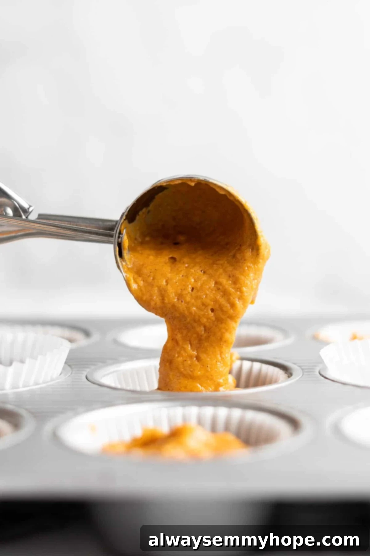 An ice cream scoop carefully depositing a portion of creamy pumpkin cupcake batter into a cupcake liner nested within a cupcake pan, ensuring even distribution.