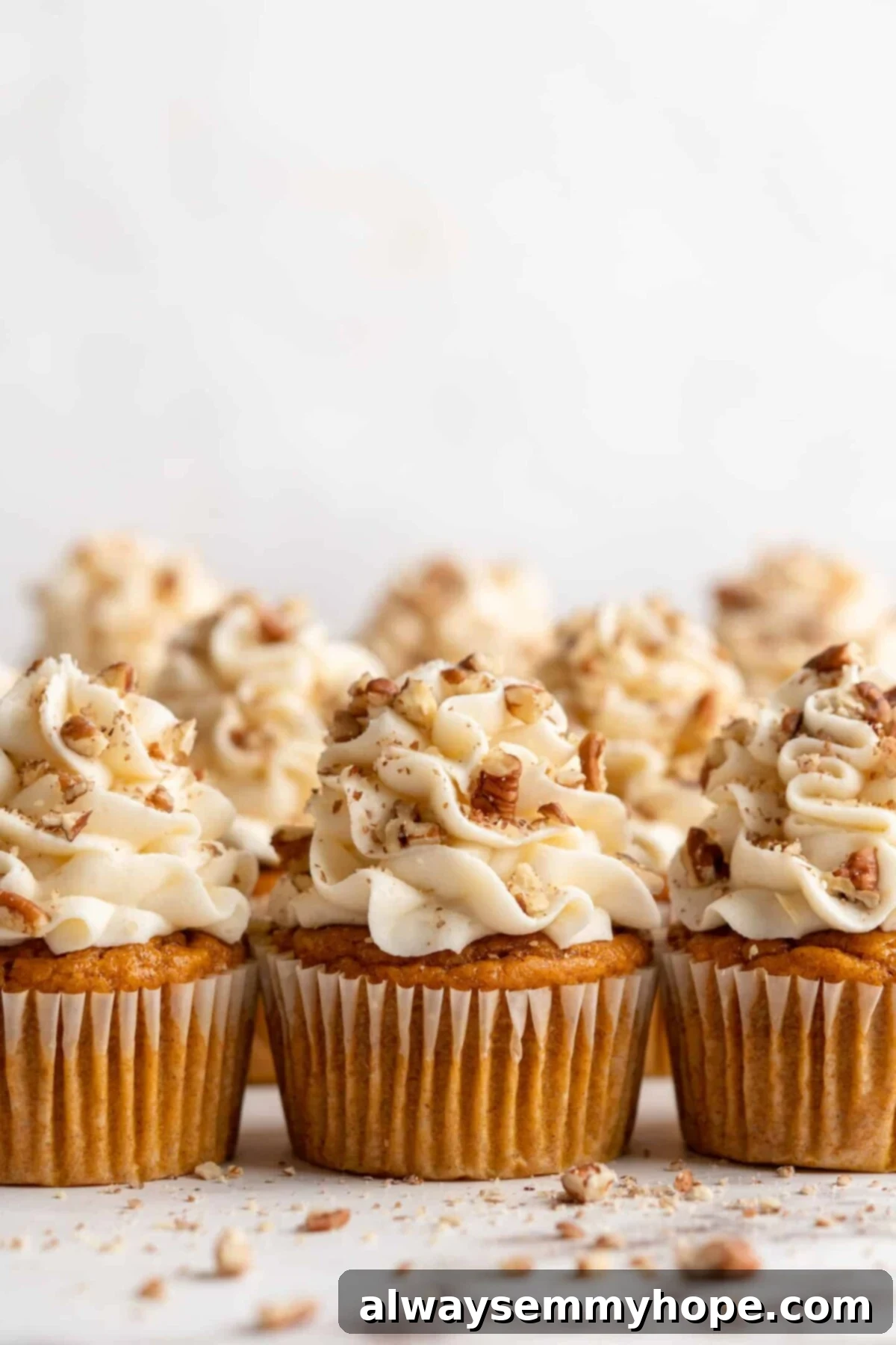 A trio of perfectly frosted vegan pumpkin cupcakes adorned with cream cheese frosting and a sprinkling of chopped pecans, with three more delicious cupcakes enticingly blurred in the background.