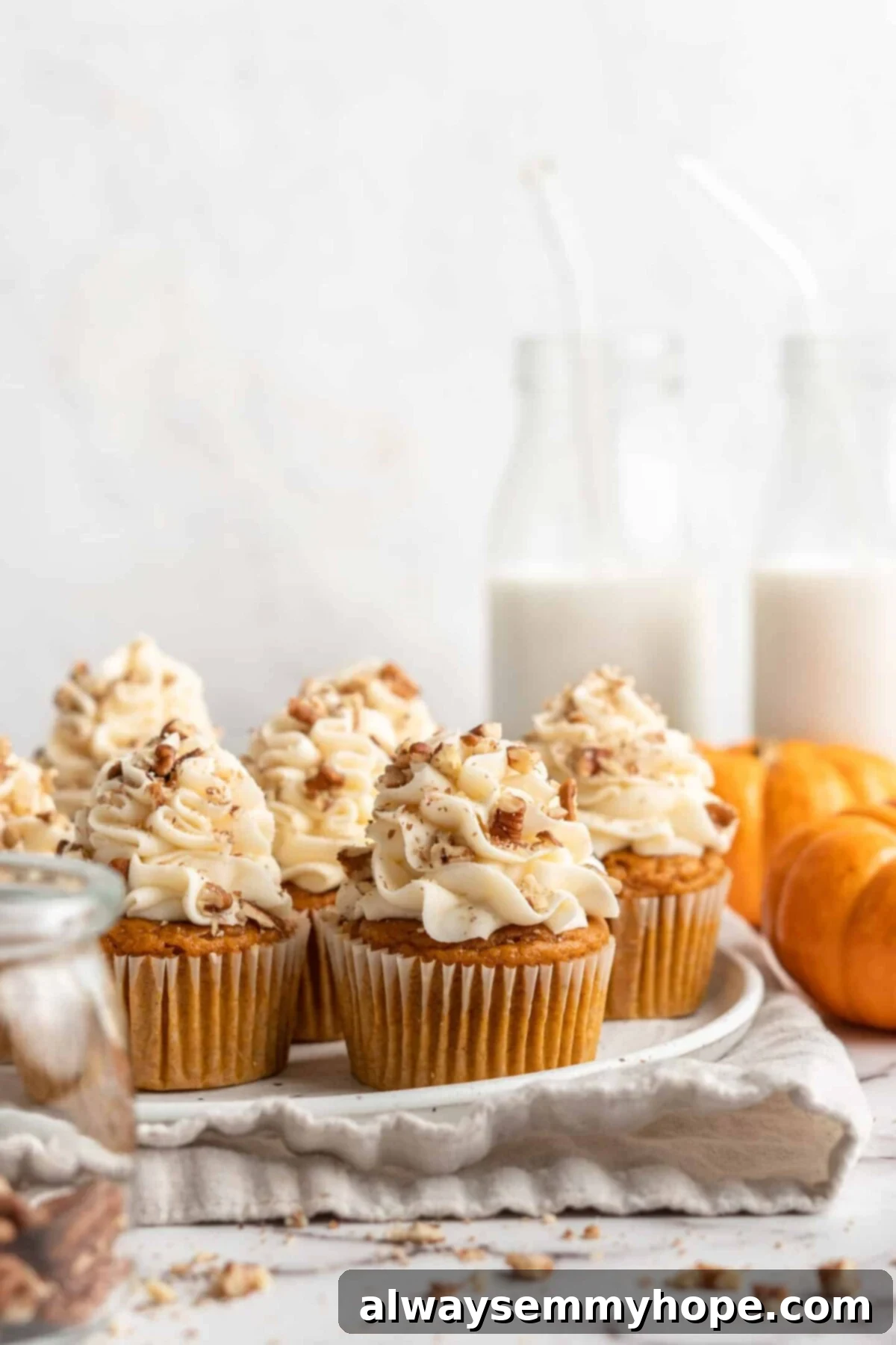 A beautifully arranged plate filled with several vegan pumpkin cupcakes, generously topped with cream cheese frosting and crushed pecans. In the background, decorative pumpkins and glasses of milk create a cozy autumnal scene.