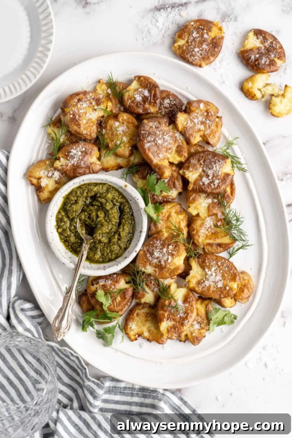 This air fryer smashed potatoes recipe is salty, crispy on the outside, and light and fluffy on the inside. It's an easy side dish, and vegan! Overhead view of a tray of smashed potatoes garnished with vegan cheese and parsley, with a bowl of pesto with a spoon in it