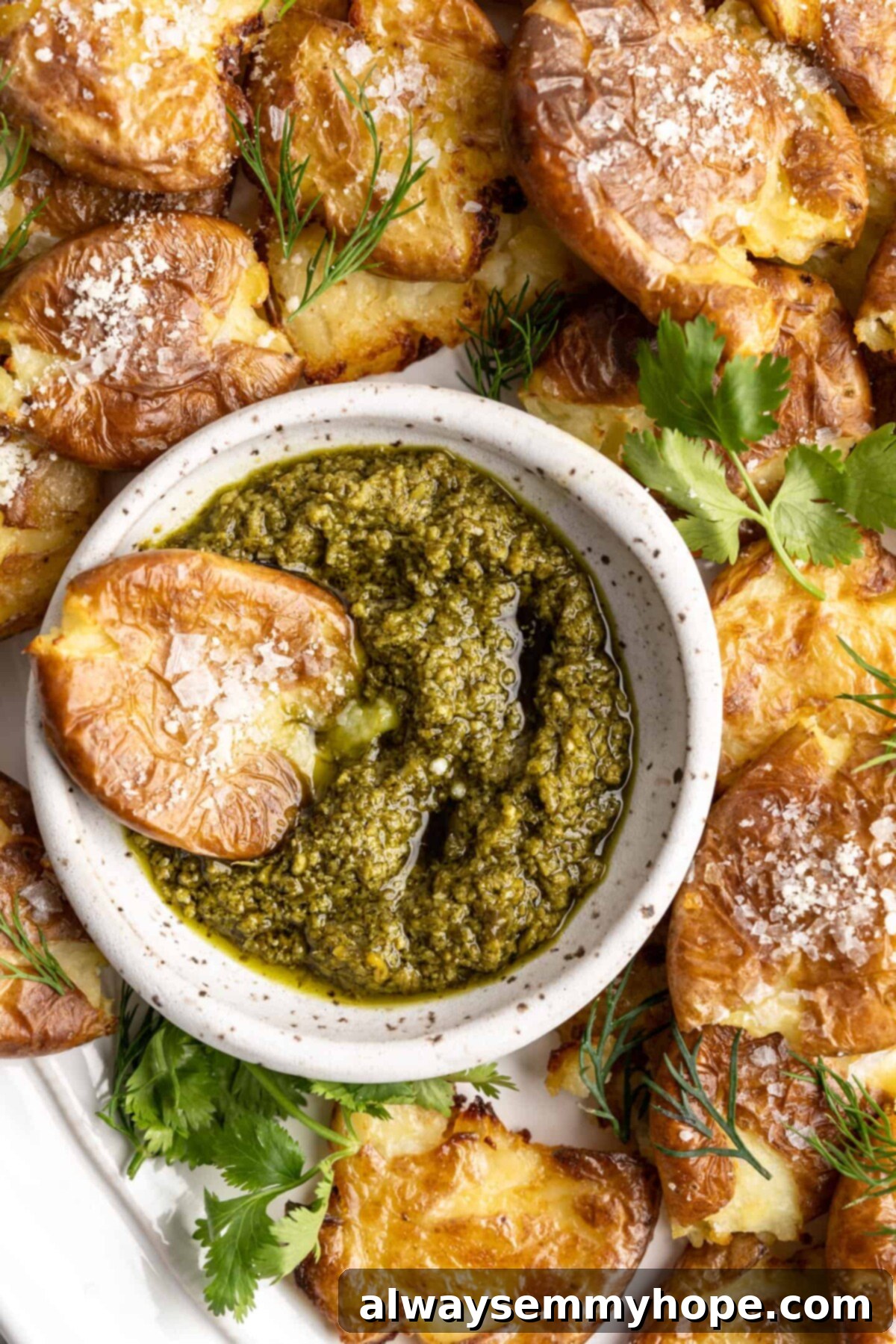 This air fryer smashed potatoes recipe is salty, crispy on the outside, and light and fluffy on the inside. It's an easy side dish, and vegan! A smashed potato resting in a bowl of pesto, on a plate full of smashed potatoes