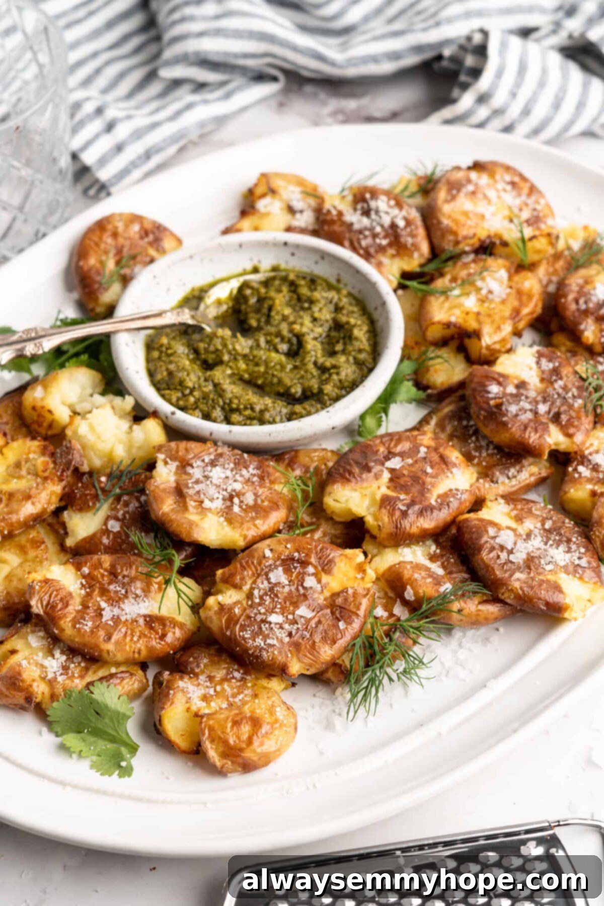 This air fryer smashed potatoes recipe is salty, crispy on the outside, and light and fluffy on the inside. It's an easy side dish, and vegan! A tray full of smashed potatoes, topped with cheese, with a bowl of pesto with a spoon in it