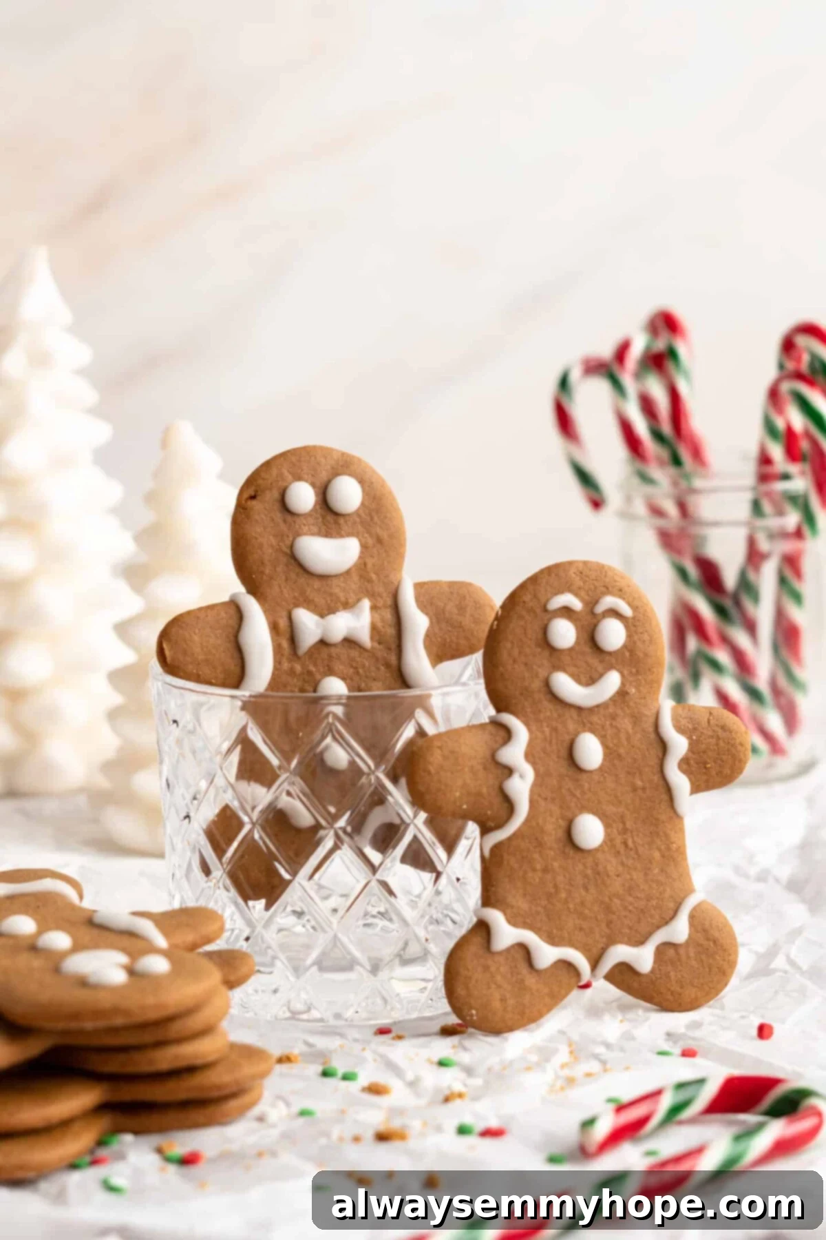 Two gingerbread men standing upright, one in a glass cup, next to a stack of gingerbread men, with a cup of candy canes in the background