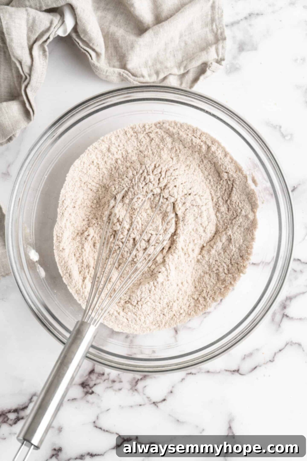 A glass mixing bowl full of flour and spices, with a whisk