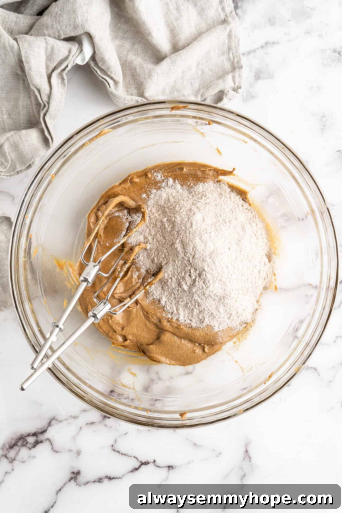 A glass mixing bowl with the wet ingredients for gingerbread men dough beat together, with a pile of dry ingredients on top, and two electric beater blades in the bowl