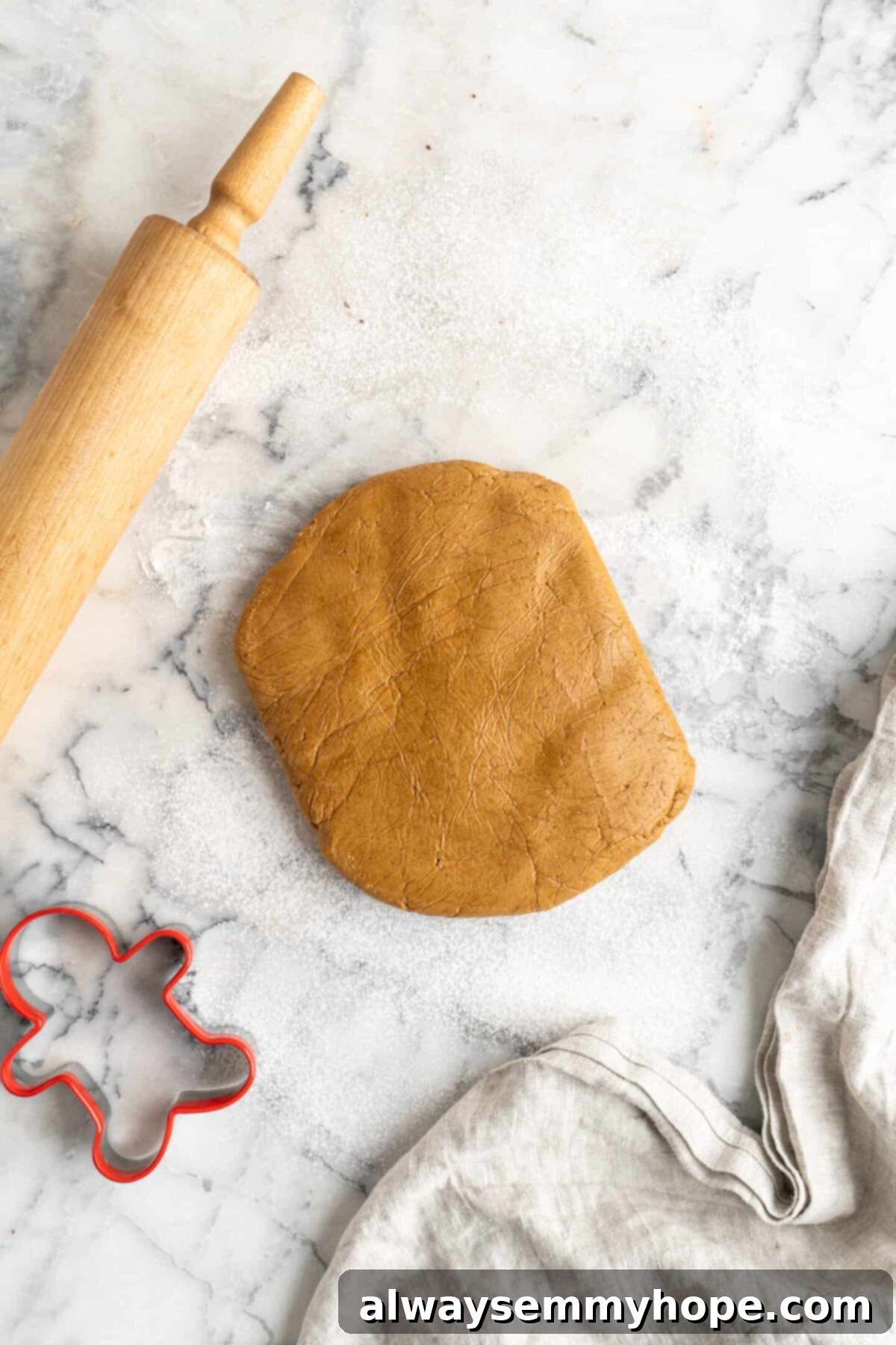 A think disk of gingerbread men dough on a marble counter, next to a kitchen towel, a gingerbread men cookie cutter, and a rolling pin
