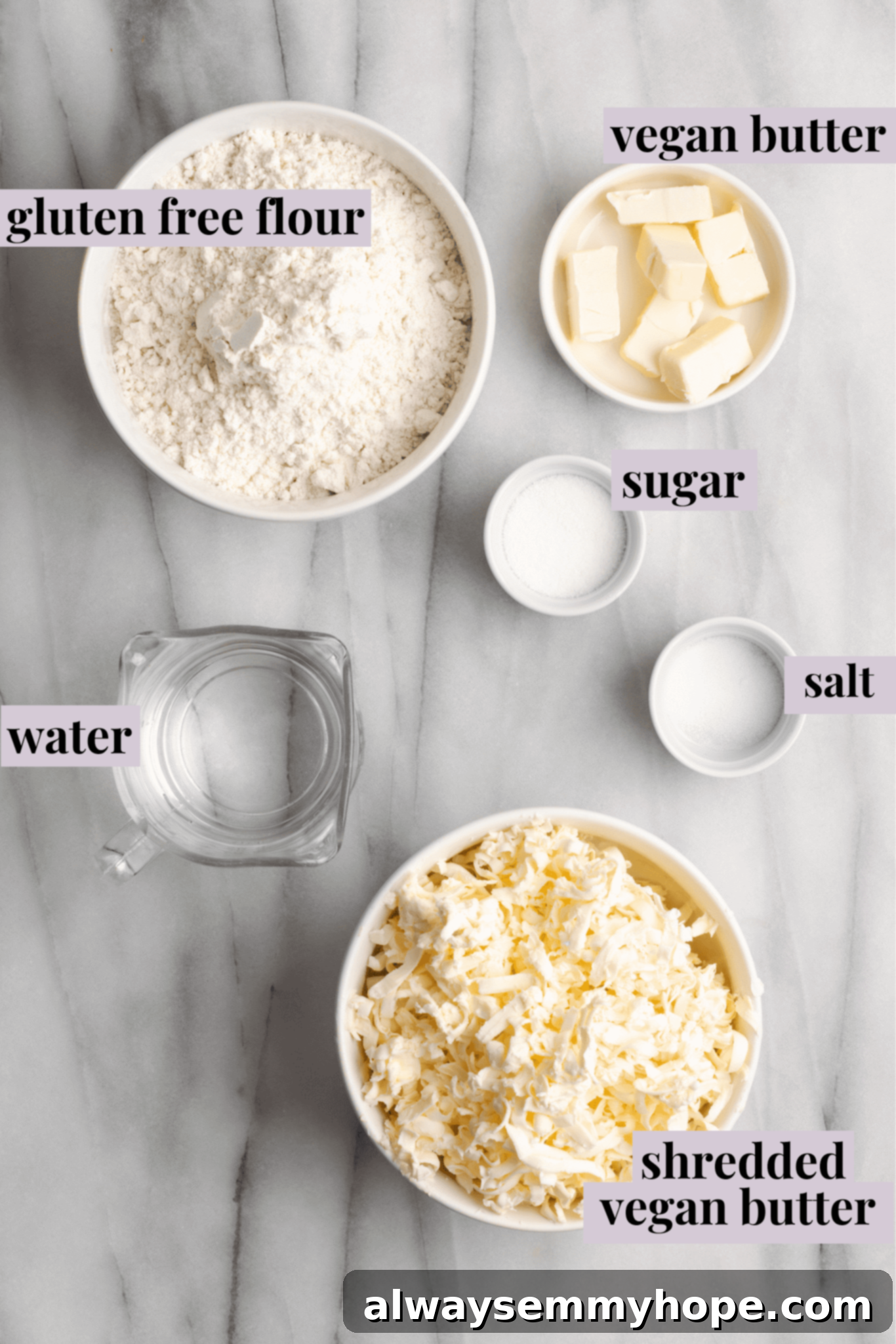 Overhead view of the ingredients for vegan puff pastry: a bowl of gluten-free dough, a glass of water, a bowl of salt, a bowl of sugar, a bowl of cubed vegan butter, and a bowl of shredded vegan butter