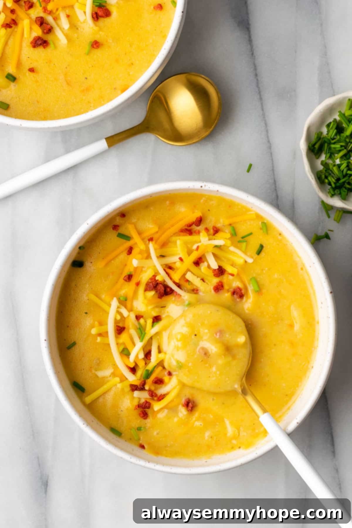This creamy vegan Instant Pot potato soup recipe is hearty, comforting, and perfect for winter, plus it's easy and quick to make! A comforting tableau: a bowl of creamy potato soup adorned with chives, vegan bacon bits, and vegan cheese, with a spoon taking a scoop, positioned next to another serving bowl and a spoon, suggesting a shared meal.