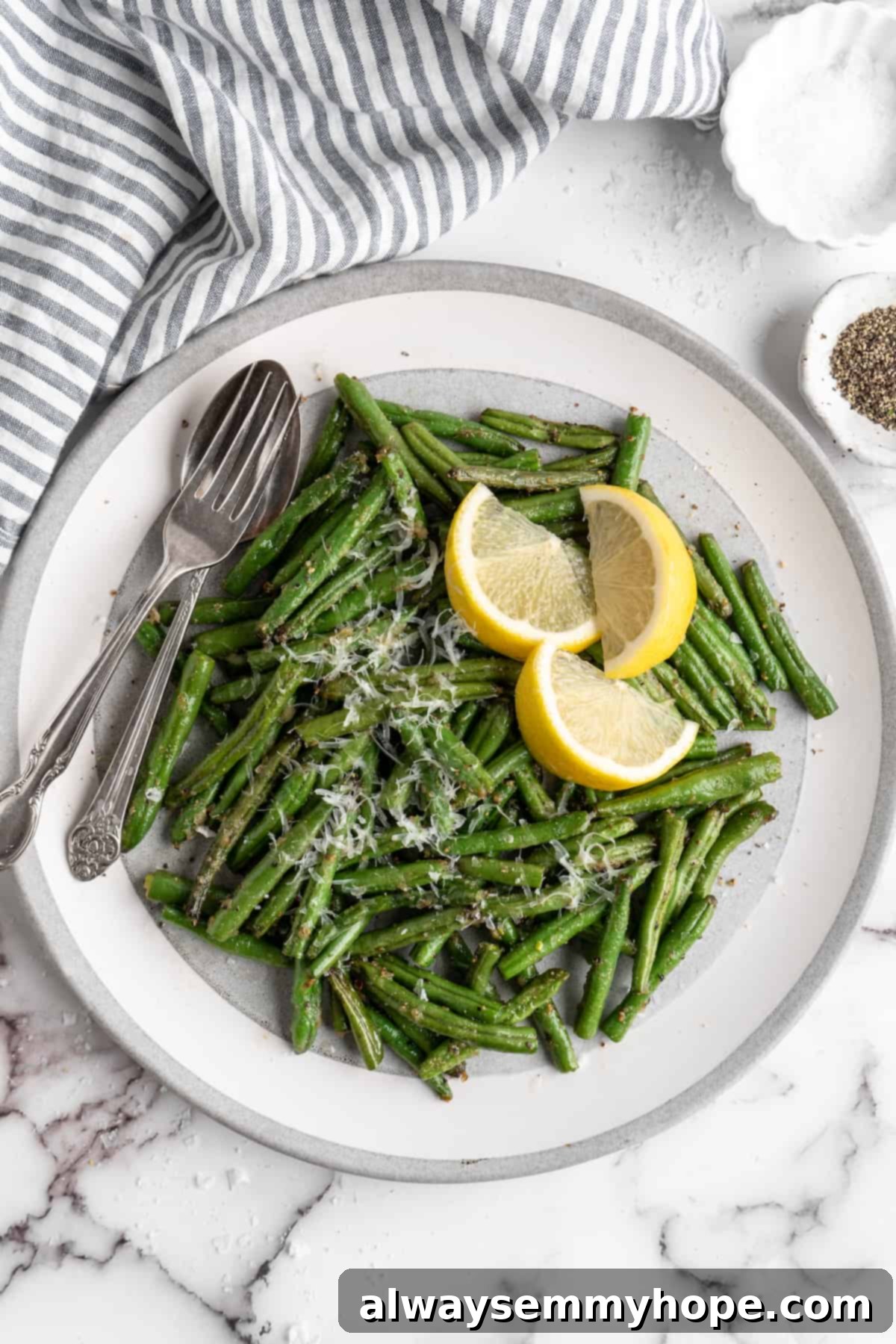 These air fryer green beans are seasoned to perfection and air fried until crispy for an easy side dish that goes with anything! Overhead view of perfectly cooked air fryer green beans on a white plate with a spoon, fork, and fresh lemon wedges, ready to serve.