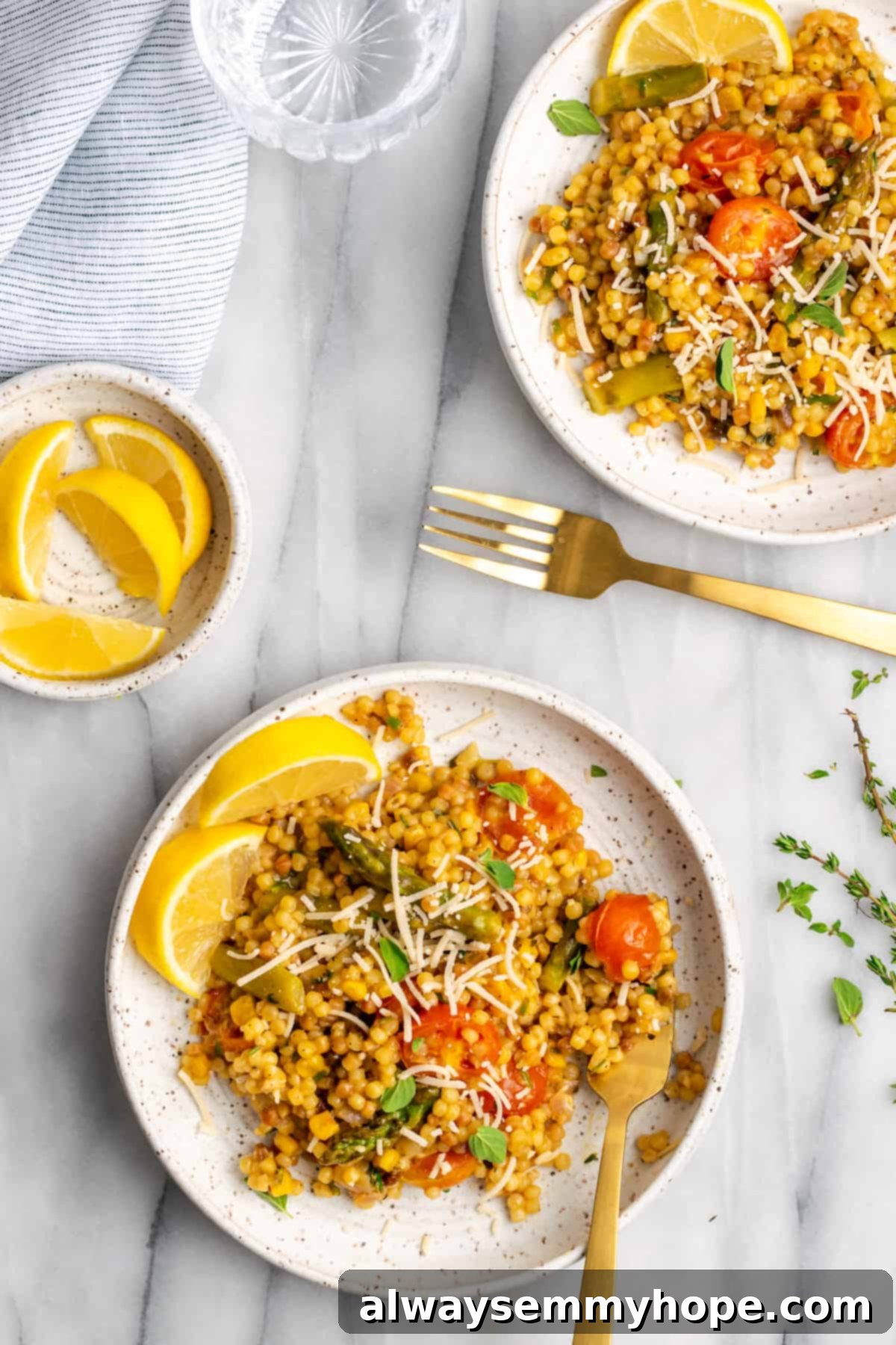 Two plates of garnished Herb & Veggie Fregola served side-by-side with a small bowl of lemon wedges, highlighting its vibrant presentation.
