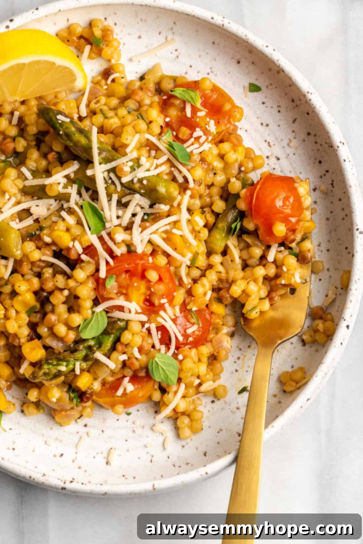 Vegan Fregola with Herbs & Veggies – A Quick & Flavorful Meal. Overhead view of a single serving of vegan fregola pasta, topped with fresh herbs and a wedge of lemon, on a white plate with a fork, emphasizing its delightful texture and appetizing presentation.