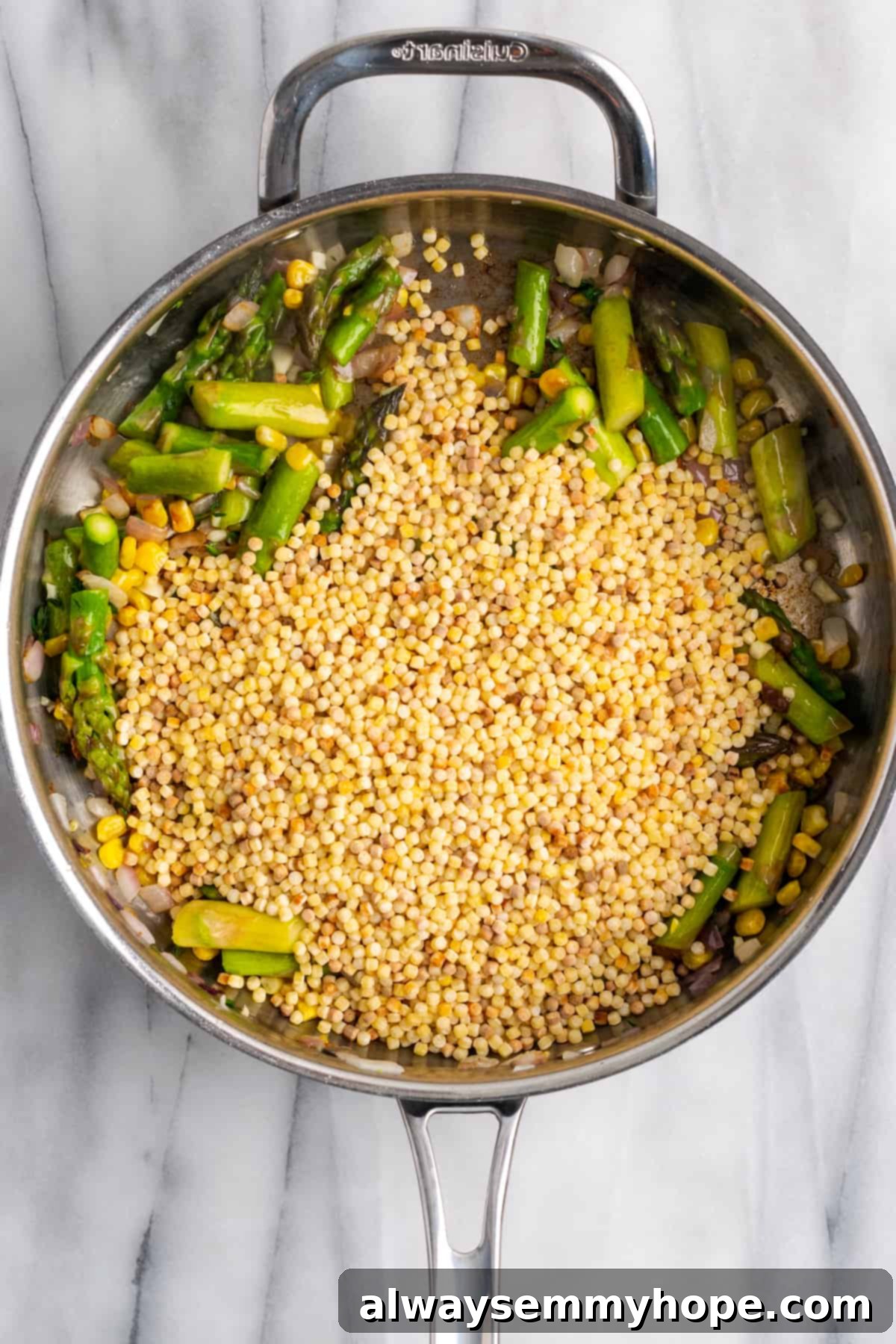 Adding Fregola to Sautéed Vegetables. Uncooked fregola sarda pasta being added to a skillet filled with sautéed vegetables like asparagus, corn, and onions, preparing for toasting.
