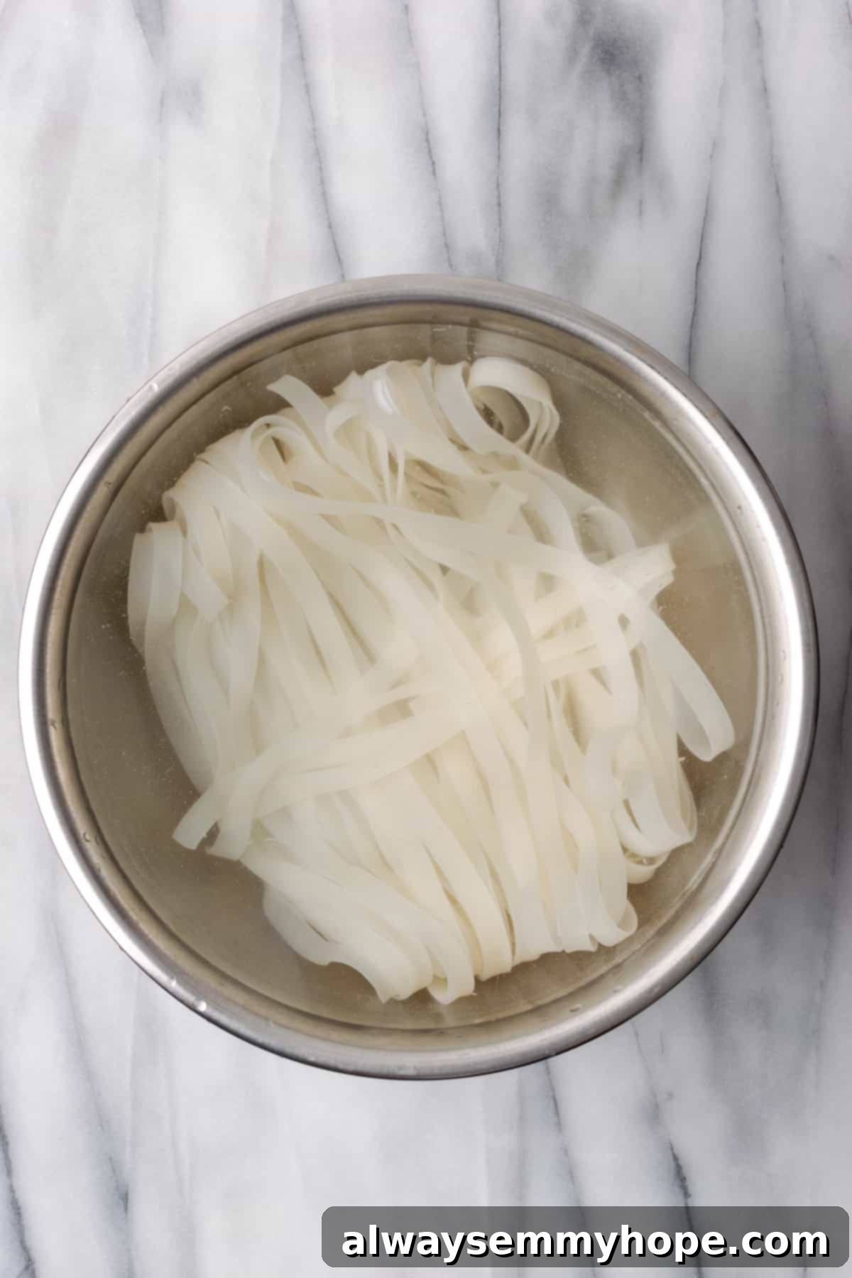 Prepping the noodles: Wide rice noodles soaking in warm water for the best vegan Chow Fun. Overhead view of XL wide rice noodles soaking in a bowl of warm water.