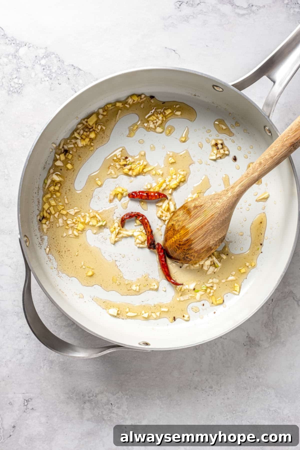 This vegan pad see ew pairs rice noodles, vegetables, and tofu with a sweet, savoury, and slightly tangy sauce for a restaurant-style meal! Overhead view of chopped garlic, ginger, and whole dried red chilis sizzling in a skillet with oil, releasing their fragrance.