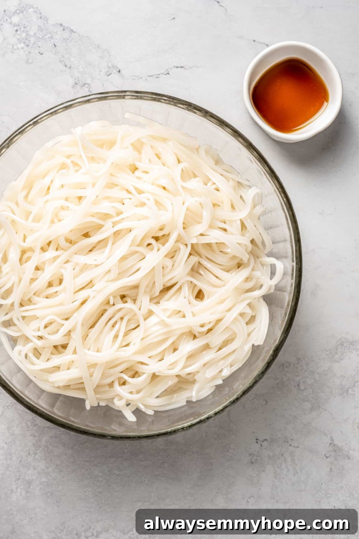 This vegan pad see ew pairs rice noodles, vegetables, and tofu with a sweet, savoury, and slightly tangy sauce for a restaurant-style meal! Overhead view of drained rice noodles in a glass bowl, with a small bowl of sesame oil beside them, ready to be tossed.