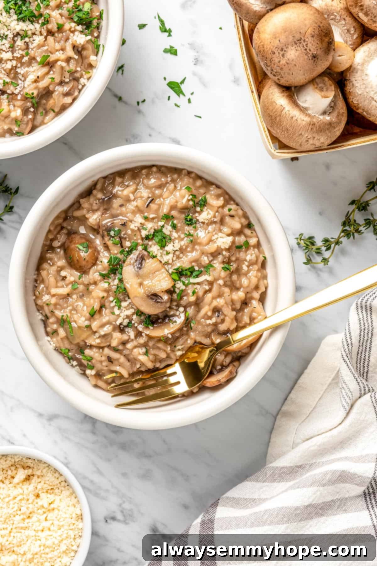 Overhead flat lay of a bowl of vegan risotto with a carton of fresh mushrooms, a patterned tea towel, herbs, and a small bowl of vegan parmesan cheese arranged on a wooden surface