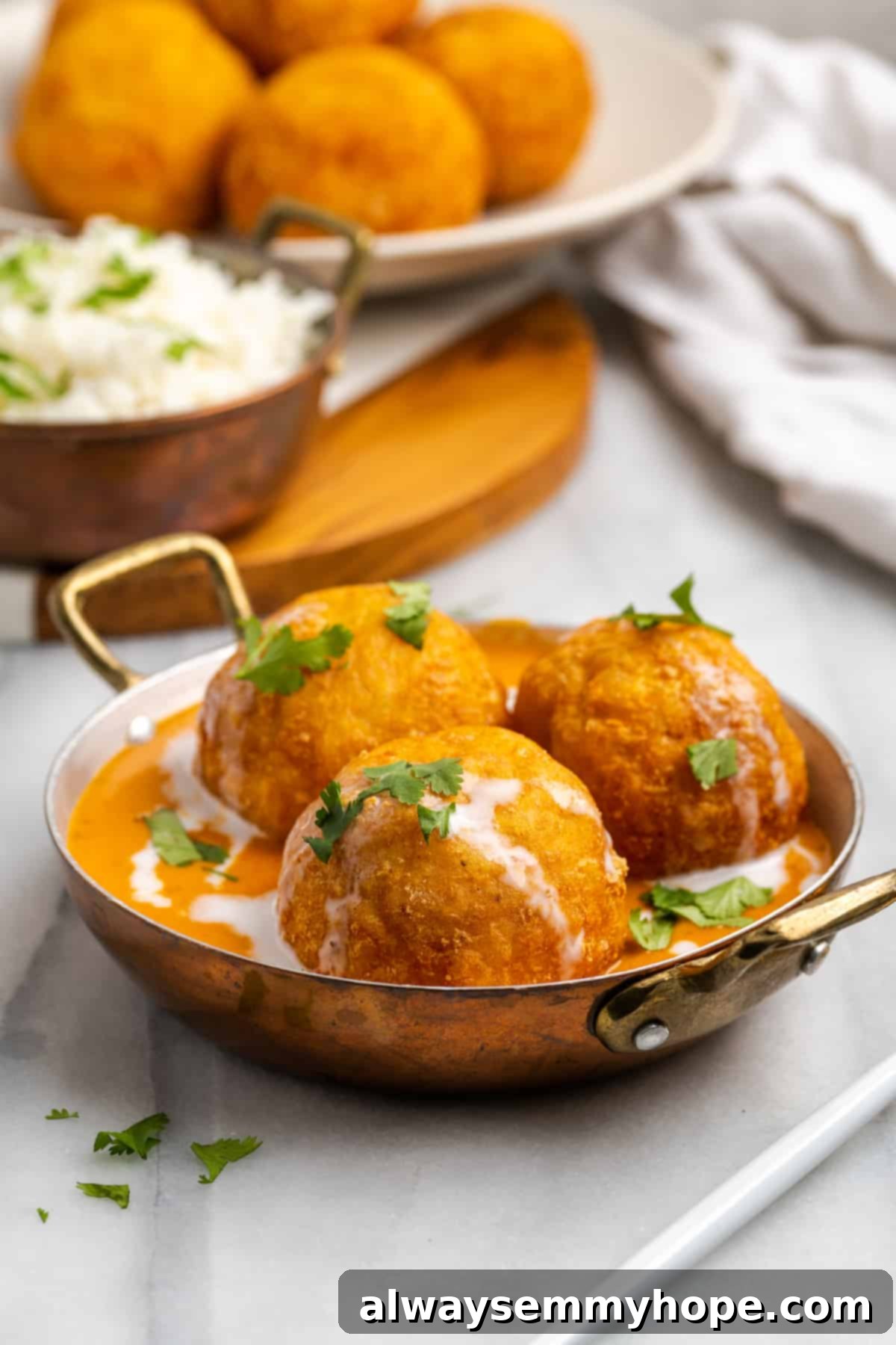 Serving bowl with 3 vegan malai kofta dumplings, with additional dumplings and rice in background