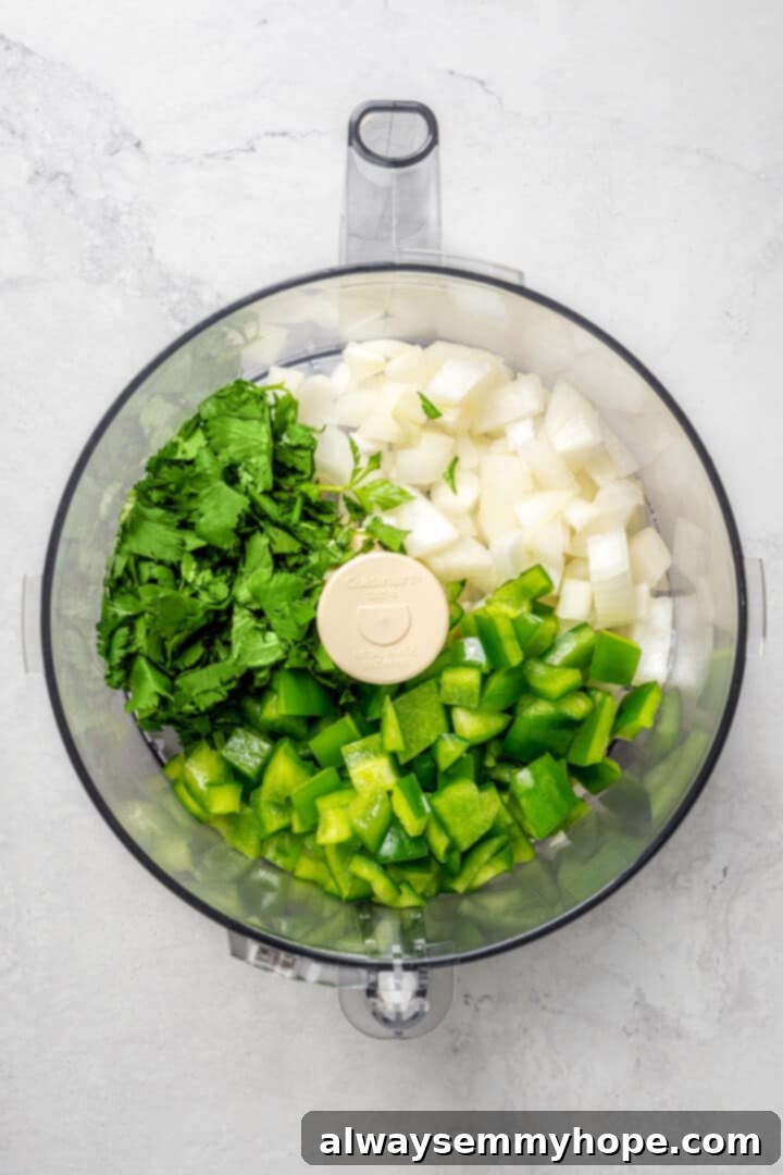 Overhead view of fresh cilantro, green bell pepper, and yellow onion in a food processor, ready to be blended for sofrito.