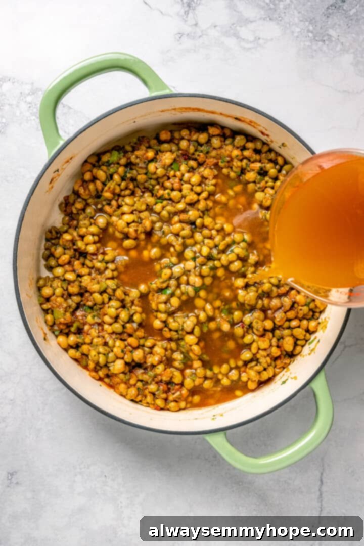 Overhead view of vegetable broth being poured into the Dutch oven, combining with the sofrito, peas, and spices before the rice is added.