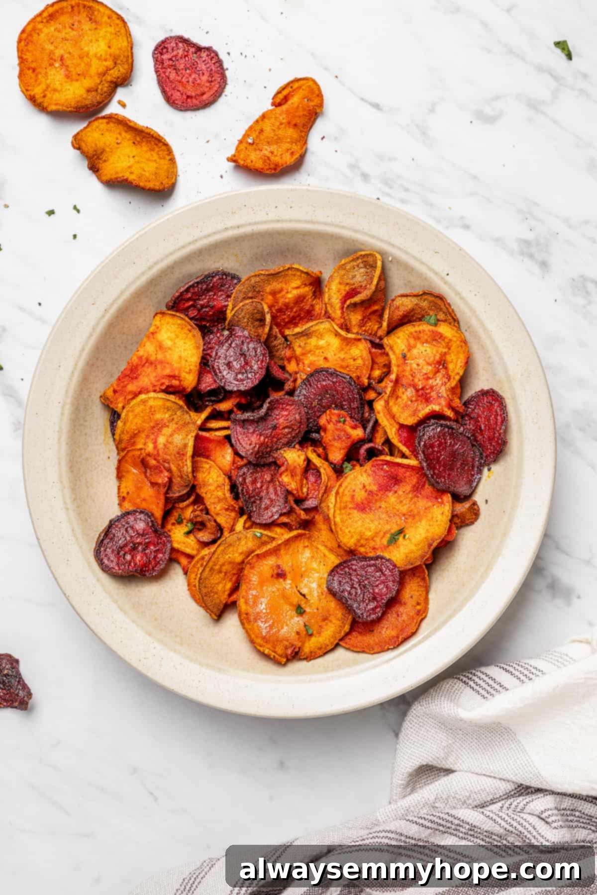 An overhead view of a shallow bowl brimming with crispy air fryer veggie chips, with more chips scattered artfully across a pristine marble countertop.