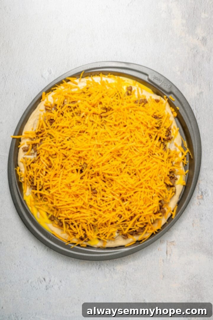 This vegan breakfast pizza features plant-based eggs, cheese, hash browns, and sausage, all baked on a crispy, chewy crust. So good! The fully assembled, unbaked vegan breakfast pizza on a round pizza pan, topped with all ingredients including a generous layer of vegan shredded cheese.