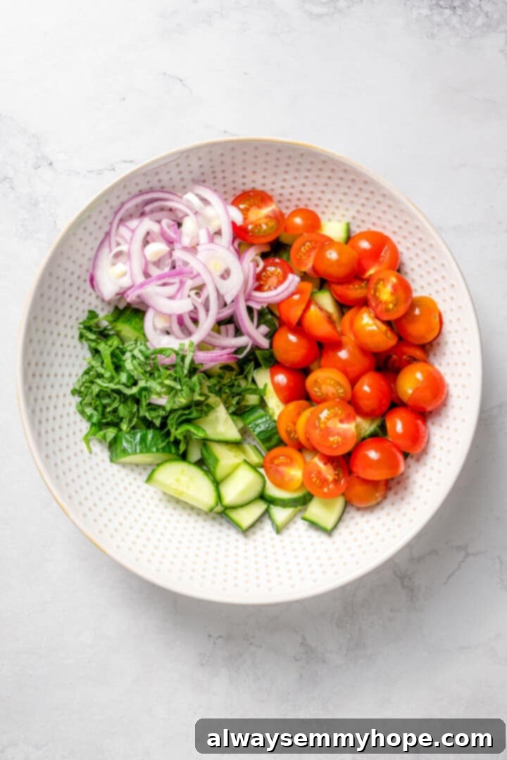 Crisp cucumbers and sun-ripened tomatoes are tossed in a simple vinaigrette for a refreshing, flavourful vegan cucumber and tomato salad. Overhead view of a bowl filled with prepped vegetables for the cucumber and tomato salad, including sliced cucumbers, halved cherry tomatoes, and thinly sliced red onion.