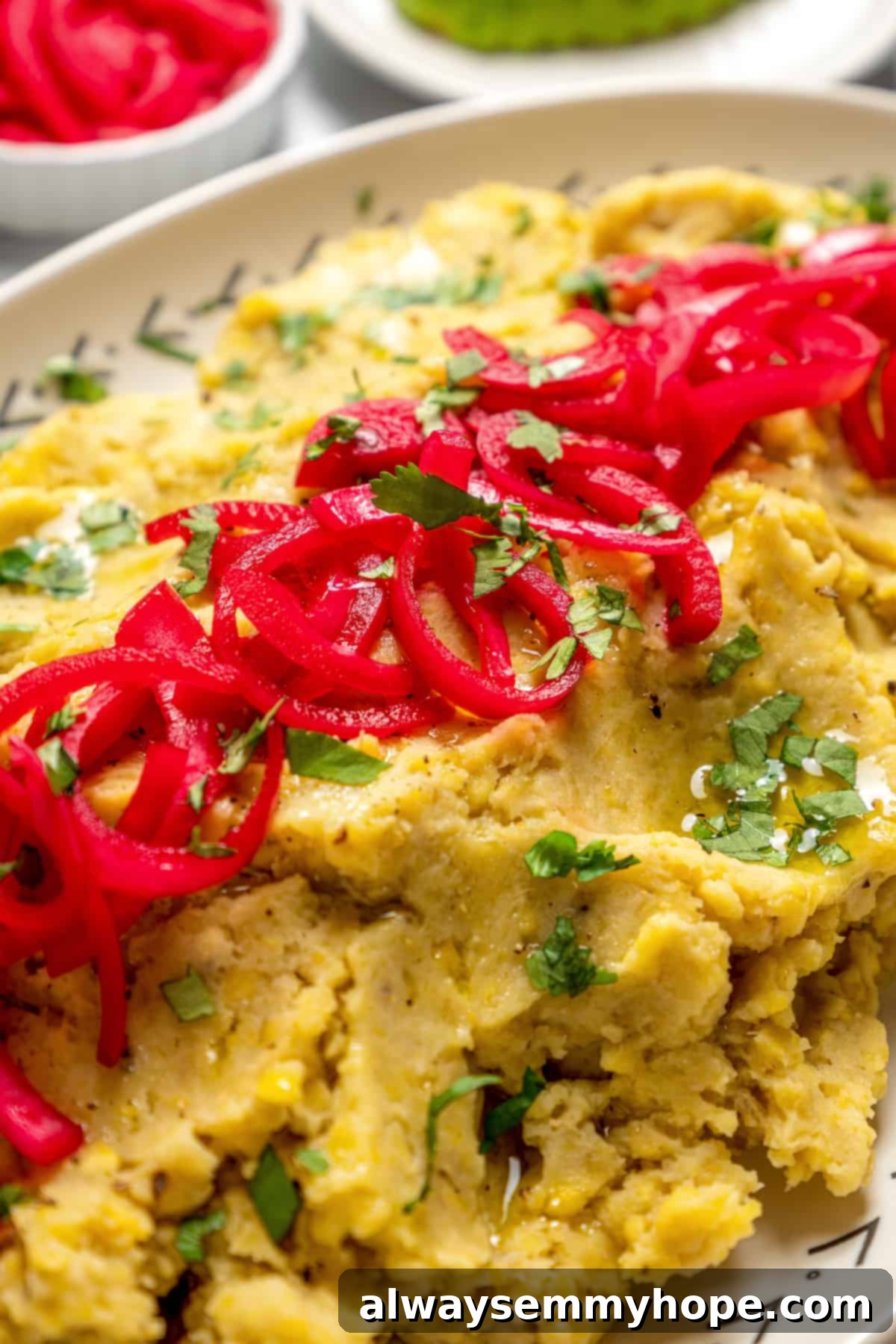 Closeup shot of creamy vegan mangú on a platter, adorned with vibrant cilantro and generous portions of bright red pickled onions.