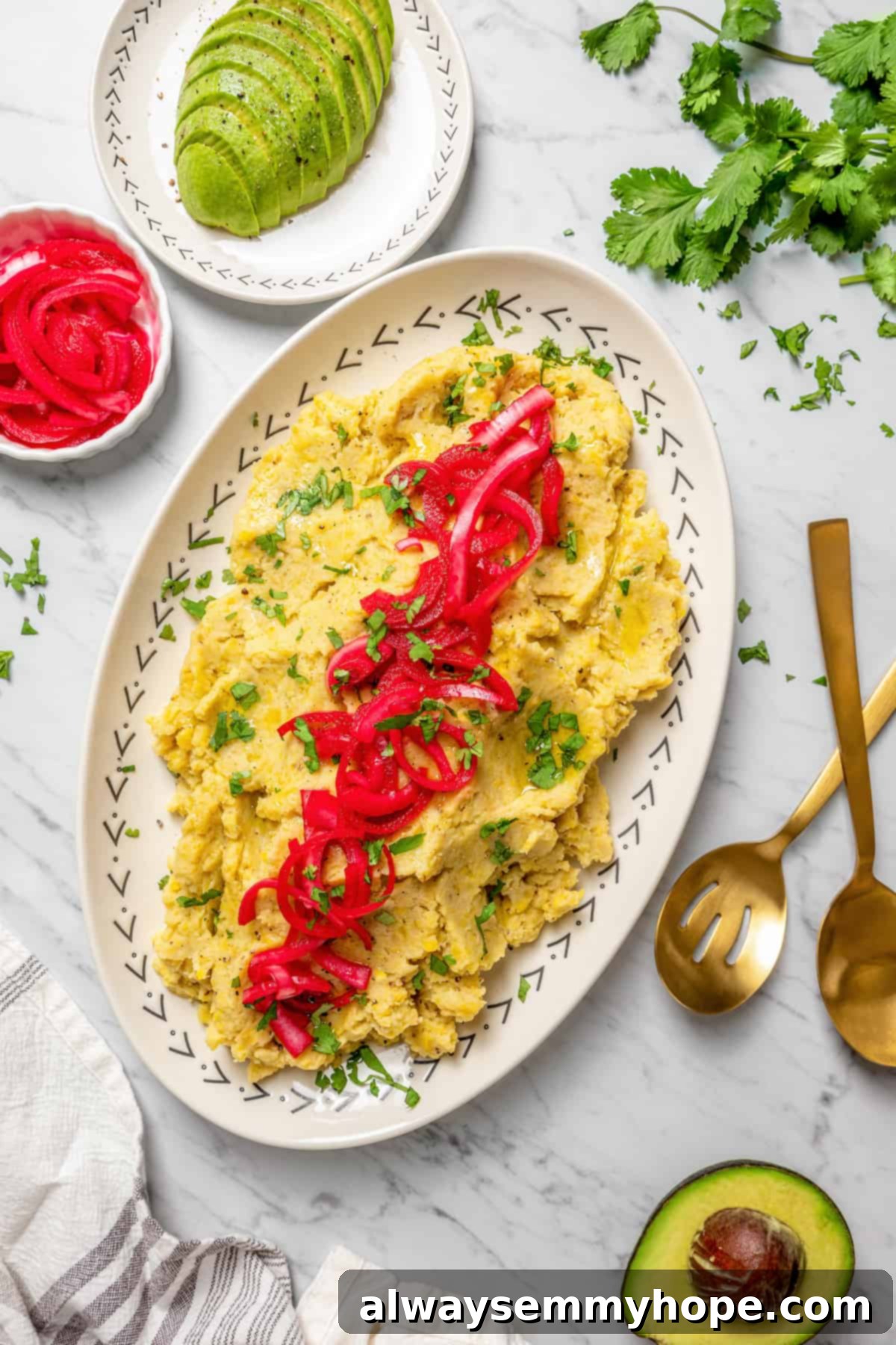 Overhead view of a central platter of mangú, artfully arranged, surrounded by small plates of fresh cilantro, creamy sliced avocado, and tangy pickled red onions.