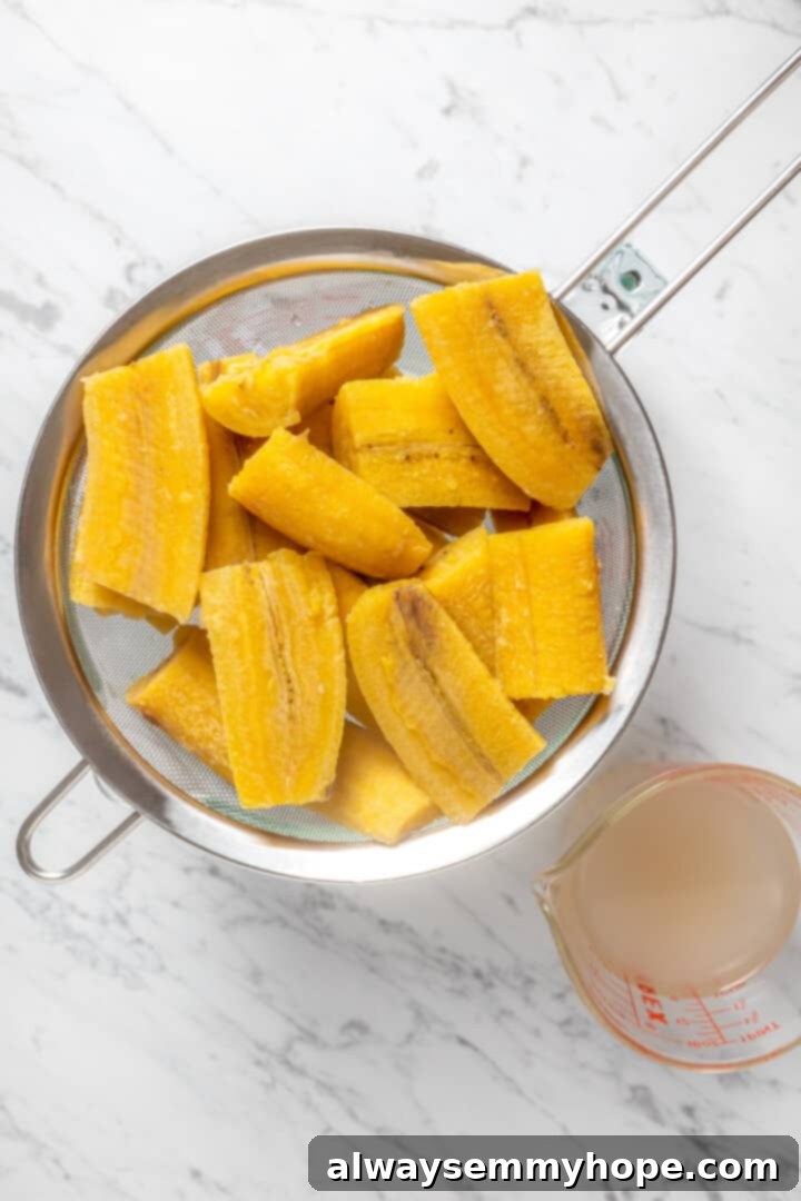 Overhead view of cooked plantains draining in a sieve, steam gently rising, after being removed from the pot.