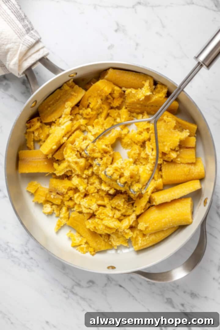 A potato masher actively mashing tender, cooked plantains directly in the pot, beginning the process of creating creamy mangú.