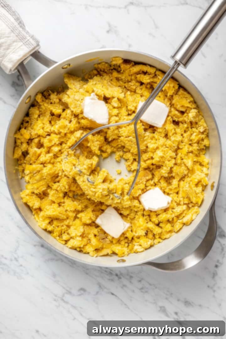 Overhead view of mashed plantain in a pot, with a potato masher resting alongside, and cubes of vegan butter melting into the mixture.