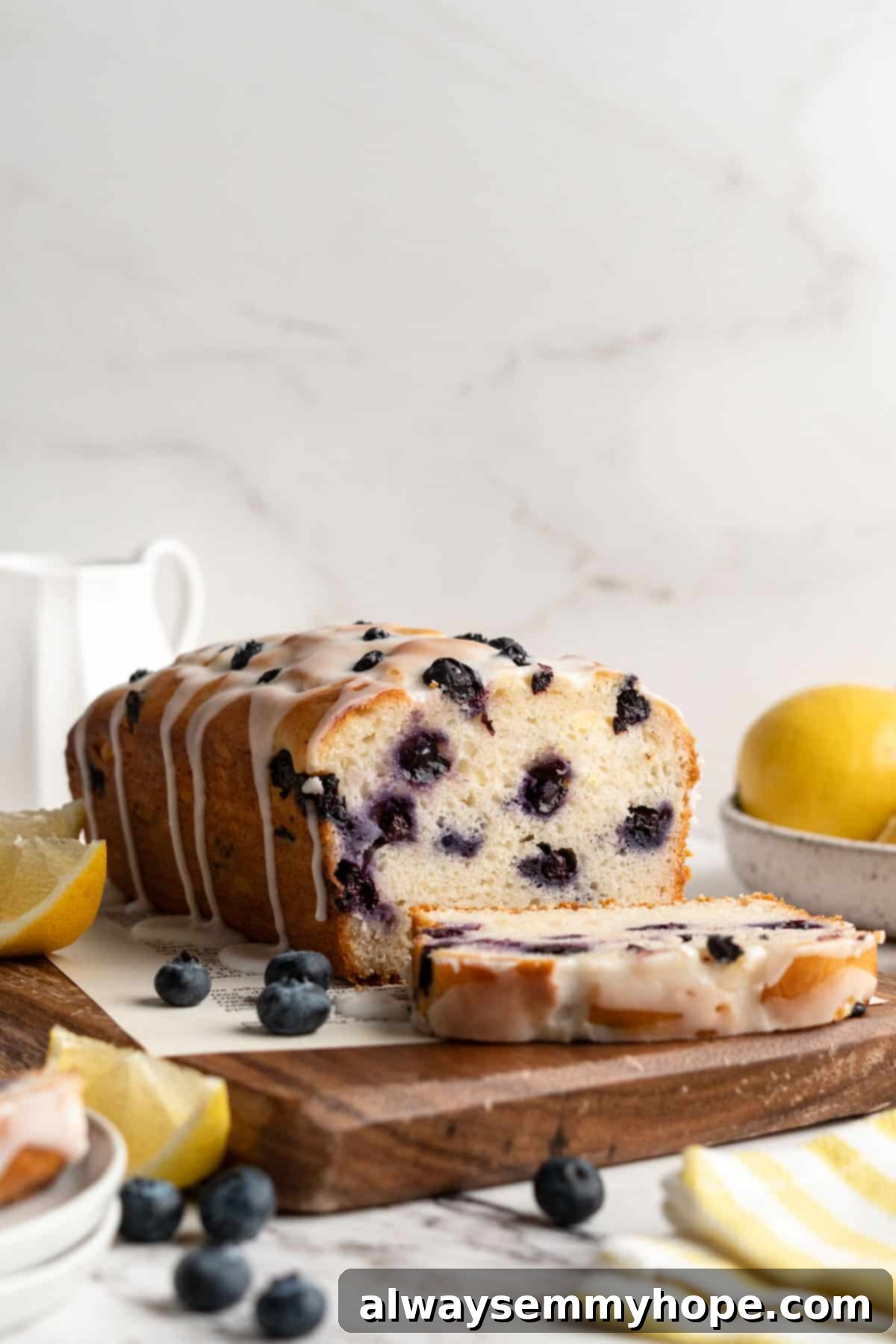 Glazed vegan lemon blueberry loaf on a wooden cutting board with one slice perfectly cut, showcasing its moist interior and beautiful glaze.