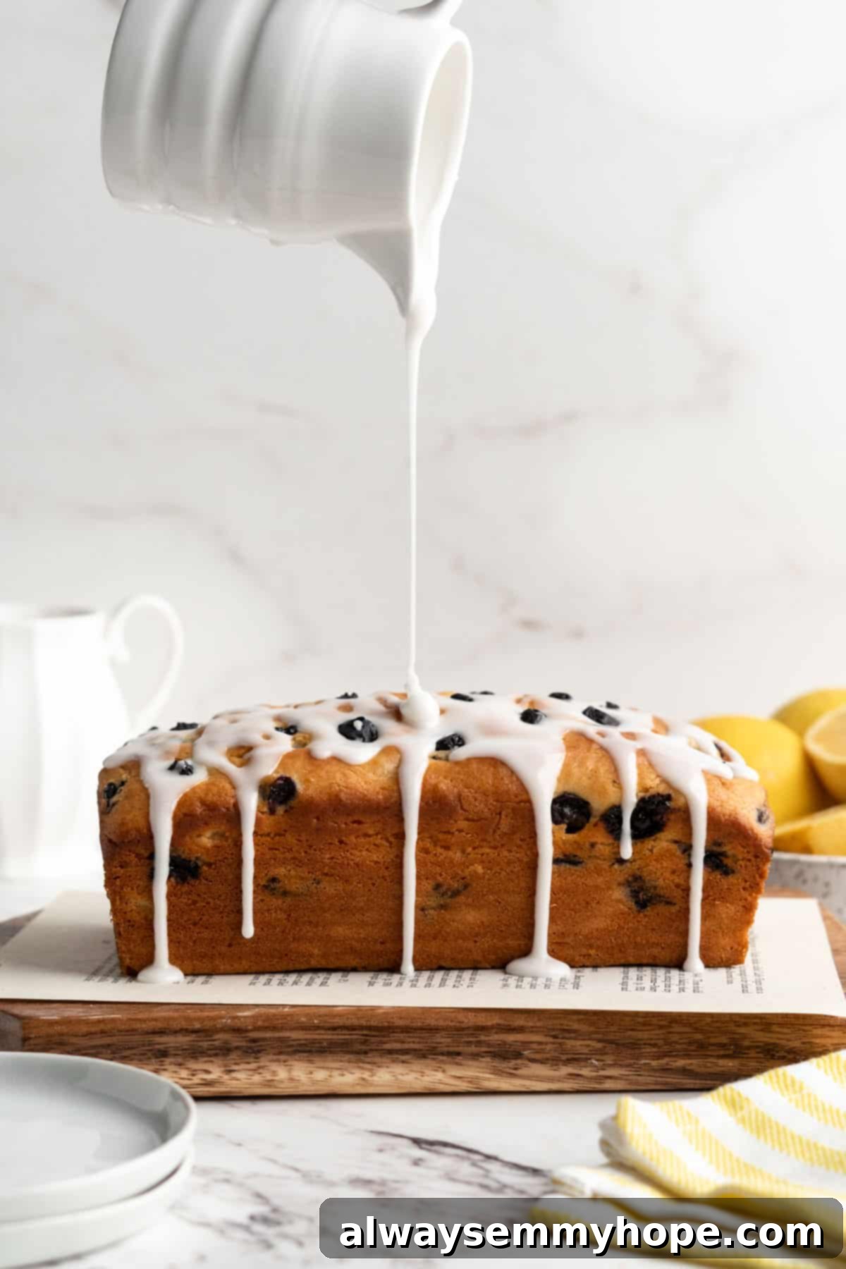Pouring a vibrant lemon glaze over the cooled vegan lemon blueberry loaf set on a wooden board.