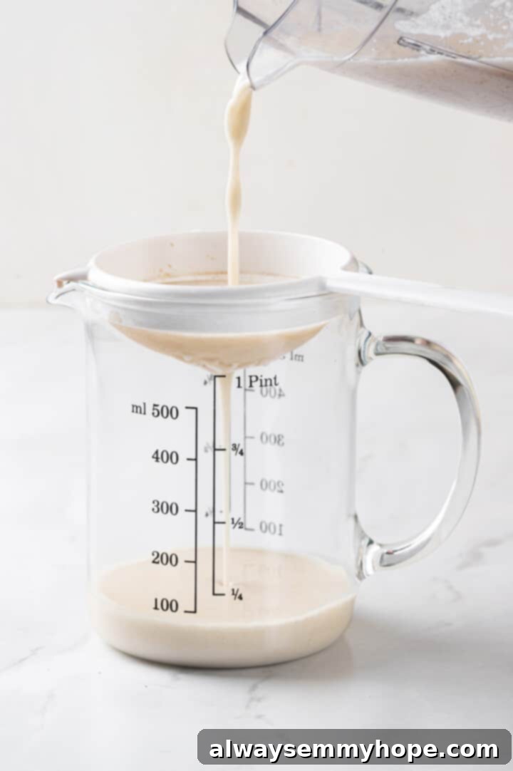 Homemade oat milk coffee creamer being poured through a fine mesh strainer into a glass measuring cup, ensuring a perfectly smooth consistency.
