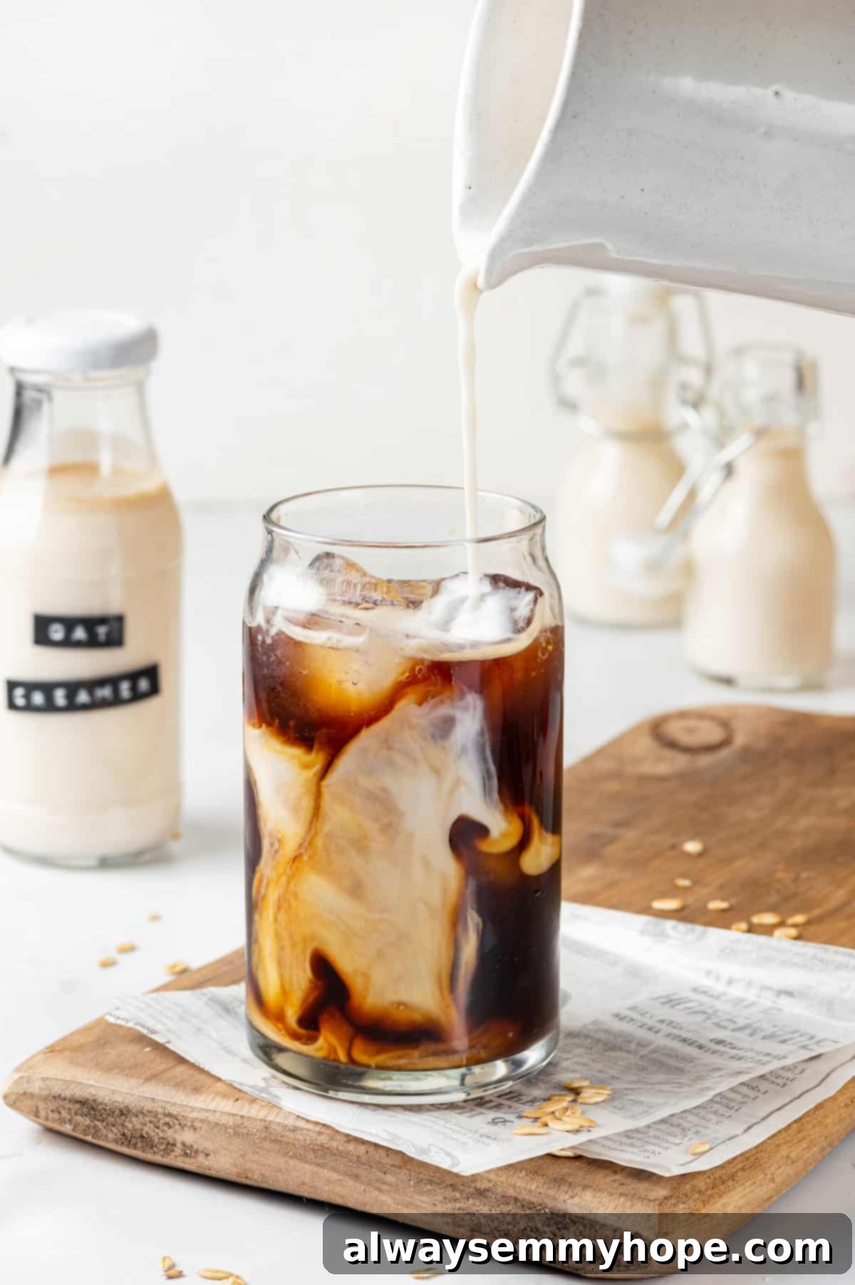 Homemade oat milk coffee creamer being poured from a pitcher into a glass of iced coffee, creating a beautiful swirl.