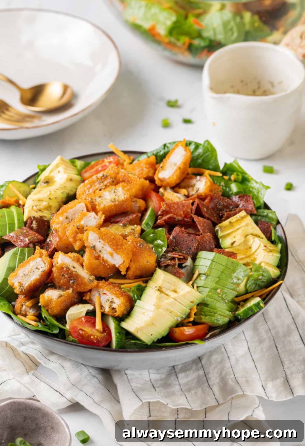 A vibrant vegan Chick-fil-A Cobb salad in a large serving bowl, with an empty salad bowl and a glass bowl of dressing in the blurred background. The salad features plant-based chicken, bacon, avocado, tomatoes, corn, and greens.