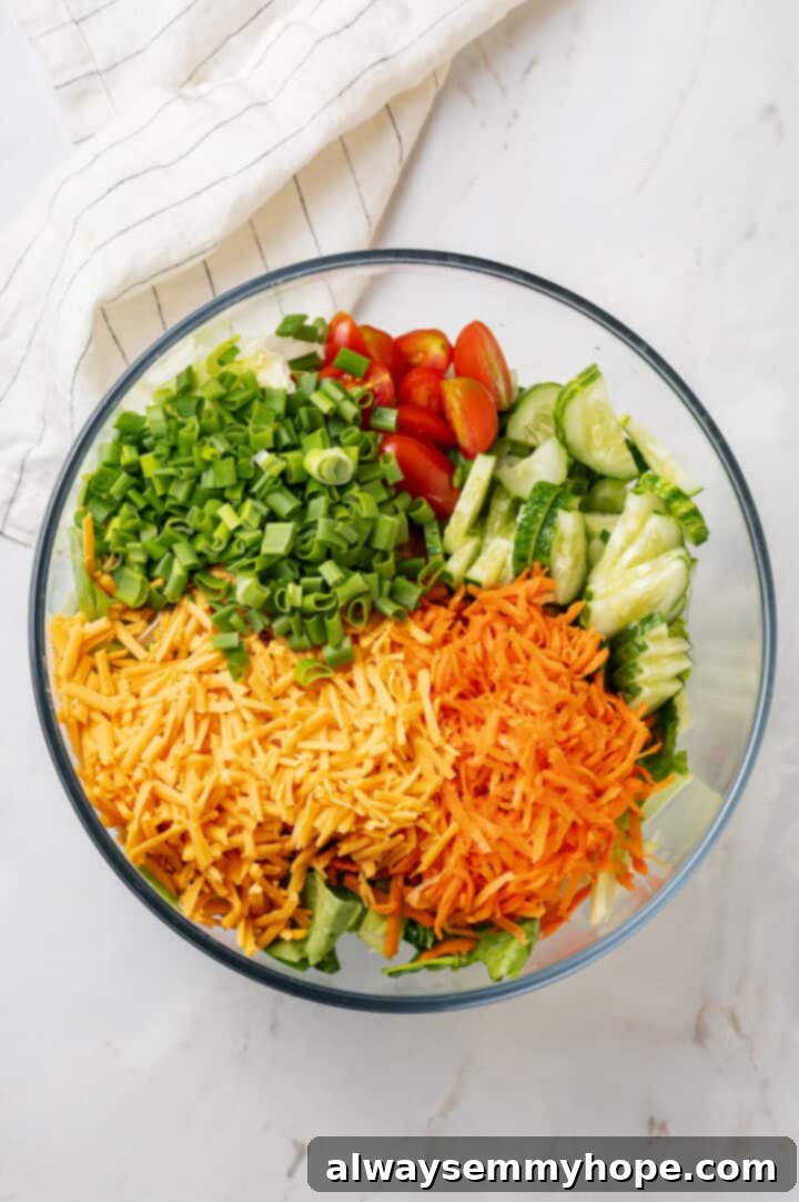 Overhead view of fresh ingredients for vegan Chick-Fil-A Cobb salad in a clear glass bowl, featuring chopped lettuce, green onions, and shredded carrots before other toppings are added.