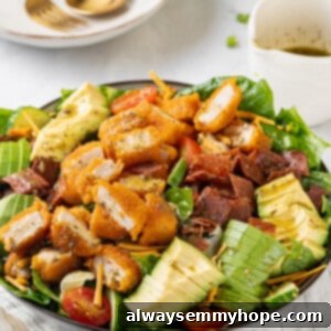 A vibrant and fresh vegan Cobb salad served in a bowl, with a fork and spoon on the side and a blurred background of kitchen elements.