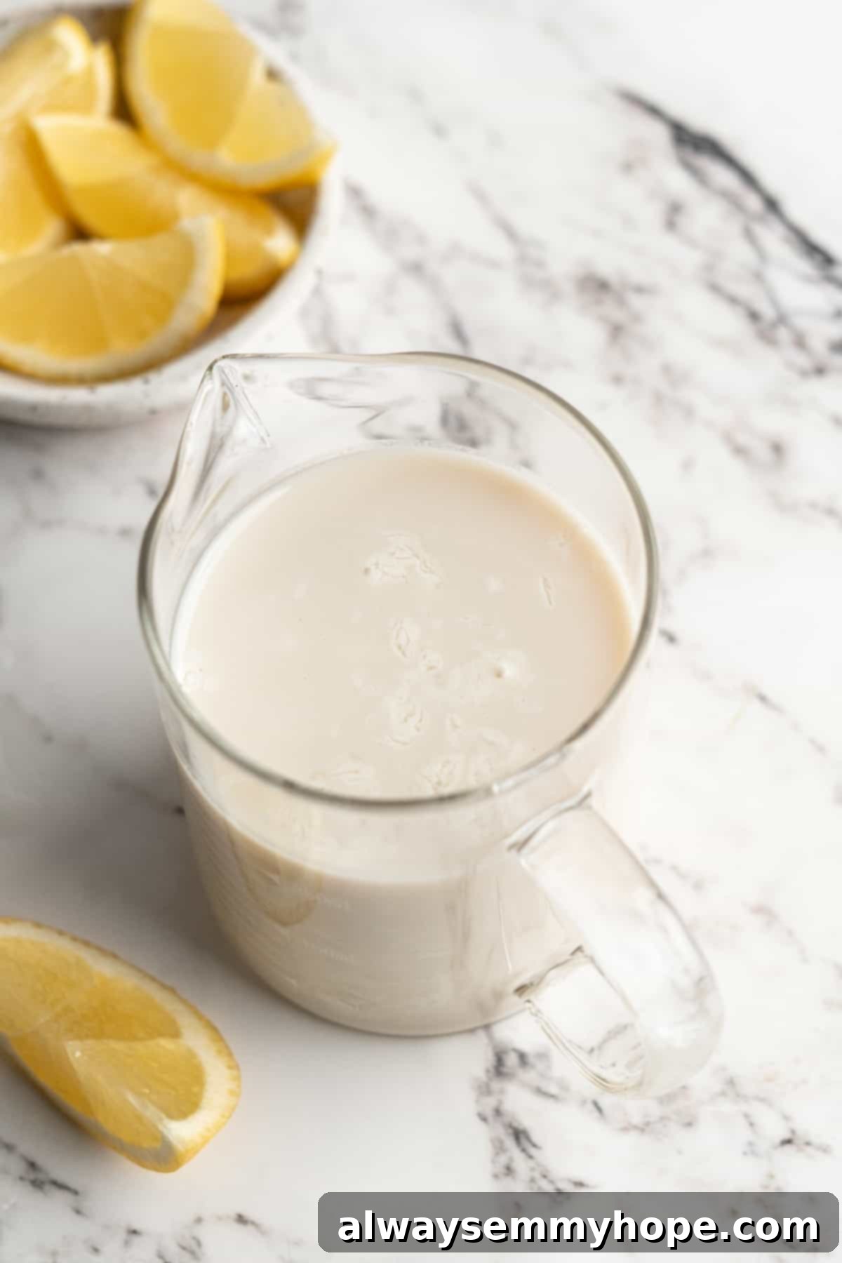 Your simple, two-ingredient vegan buttermilk is ready for all your favorite recipes! A liquid measuring cup full of prepared vegan buttermilk with a bowl of fresh lemons in the soft-focus background.